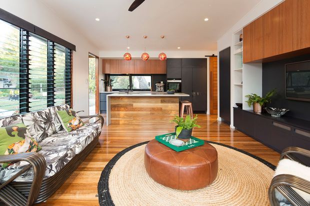 A bright open-plan living room and kitchen with warm timber flooring and cabinetry, a dark built-in media wall, black-framed louvred windows, a timber island, patterned rattan seating, and layered pendant lighting. A round leather ottoman and woven circular rug define the living area, while indoor plants add greenery.