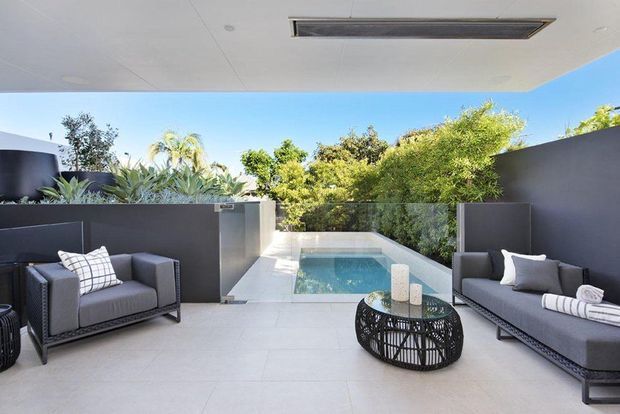 A contemporary covered outdoor lounge overlooking a compact rectangular swimming pool. The space features pale tiled flooring, dark charcoal privacy walls, glass pool barriers, grey upholstered seating, and a round black metal coffee table, with lush planting surrounding the pool area. A recessed linear ceiling light is integrated into the roof above.
