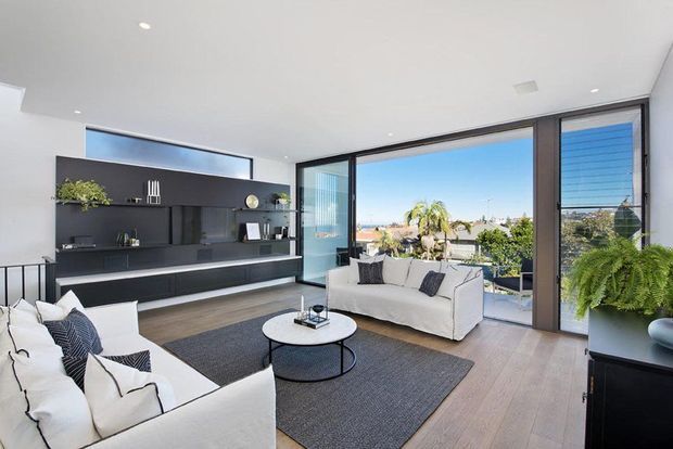 Bright contemporary living room with floor-to-ceiling sliding glass doors opening to a balcony or terrace, offering suburban and coastal views. The room features white sofas, a round coffee table, a large dark rug, pale timber flooring, and a sleek dark built-in media wall with open shelving. Recessed ceiling lights and abundant glazing create an airy, refined atmosphere.