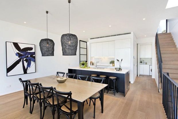 A bright open-plan dining and kitchen interior with a large light timber dining table, black timber dining chairs, a dark kitchen island with bar stools, white cabinetry, woven pendant lights, and pale timber flooring. A staircase with black balustrading rises along the right side, and contemporary artwork and greenery add visual accents.