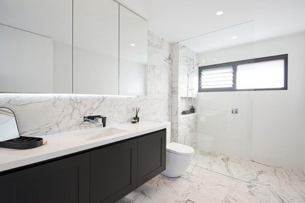 Contemporary bathroom with a long floating dark vanity, integrated white basin, mirrored cabinetry, and extensive white marble-look wall and floor tiling. A wall-hung toilet, frameless glass shower enclosure, black-framed awning window, and recessed ceiling lights complete the bright, refined space.