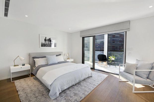 Bright, contemporary bedroom with a large upholstered bed, layered grey and white bedding, patterned area rug, timber flooring, white walls, minimalist bedside table and lamps, and a white-framed armchair. Full-height glazed doors with horizontal blinds open to a small balcony or outdoor area, bringing in abundant natural light.