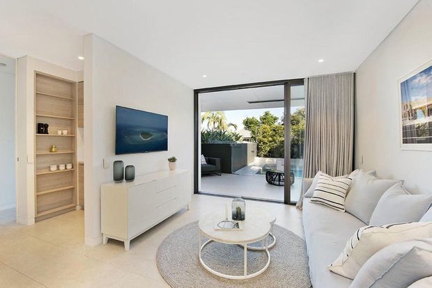 Bright contemporary living room with a white sectional sofa, round nesting coffee tables, pale rug, white media console, built-in timber shelving, and large black-framed sliding glass doors opening to an outdoor terrace. The room has a clean, airy neutral palette with warm timber accents and abundant daylight.