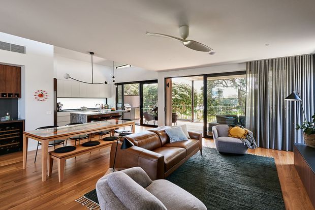 A bright, open-plan mid-century-inspired living and dining area connected to a contemporary kitchen. The room features timber flooring, a long timber dining table with bench seating, a brown leather sofa, sculptural upholstered lounge chairs, dark green rug, floor-to-ceiling glazed doors, full-height curtains, and a ceiling fan. The kitchen includes light cabinetry and a stone island, while greenery is visible beyond the glazing.