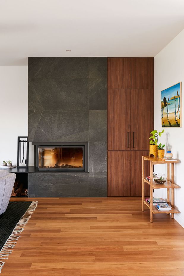 A warm contemporary living room centered on a built-in fireplace clad in large dark stone panels. The fireplace is integrated beside full-height walnut cabinetry, with timber flooring, a small open shelving unit, artwork, plants, and part of a sofa and rug visible at the edge of the frame.