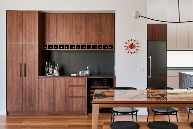 A warm contemporary open-plan dining and kitchen area with a built-in dark timber wall unit, integrated wine storage, black stone-look splashback, drawers, and concealed cabinetry. A timber dining table with a colourful patterned surface and black modern chairs occupies the foreground. The room includes timber flooring, light neutral walls, a partial kitchen visible beyond, a sculptural wall clock, and a simple globe pendant light.