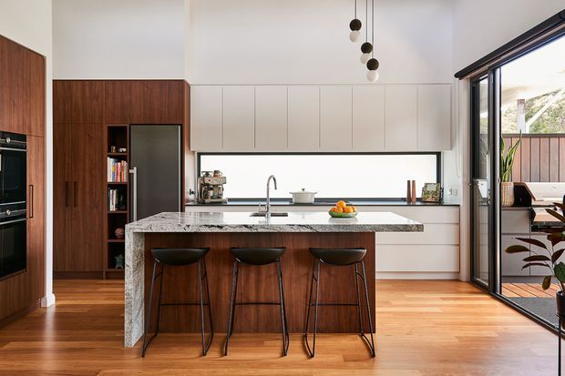 A bright, contemporary kitchen with a large stone island and three bar stools, warm timber flooring, dark timber cabinetry, and streamlined white built-in cabinetry. A wide horizontal window sits above the rear benchtop, while full-height glazed doors open to an adjacent outdoor area. Pendant lights and integrated appliances complete the refined open-plan kitchen.