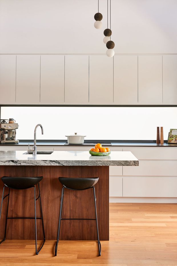 A bright, minimalist kitchen centered on a large island with a dark natural-stone benchtop and rich timber-faced cabinetry. Two black bar stools sit beneath the island overhang. White full-height cabinetry, integrated lower drawers, a slim dark splashback, stainless-steel sink and curved tap create a clean, refined composition. A sculptural cluster pendant hangs above, while timber flooring adds warmth.