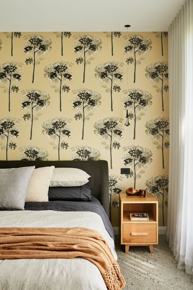 A warmly styled bedroom featuring a dark upholstered bedhead, layered white, charcoal and rust-toned bedding, and a timber bedside table. The dominant feature is a pale yellow botanical tree-patterned wallpaper, complemented by sheer full-height curtains and a terrazzo-like floor. A small pendant light and black wall switch add restrained contemporary detailing.