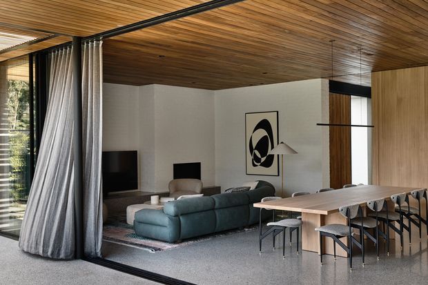 Open-plan contemporary living and dining area with a dark teal sectional sofa, compact lounge seating, an elongated timber dining table with chairs, and a linear kitchen island or counter. Full-height glazing with curtains opens to the exterior, while the room features a warm timber-lined ceiling, pale walls, polished grey flooring, black metal framing, and graphic wall art.