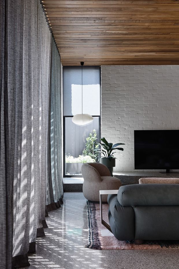 A refined living room with a dark upholstered sofa and rounded lounge chair facing a television on a white-painted exposed brick feature wall. Full-height sheer grey curtains filter daylight beside a tall window, while a warm timber slatted ceiling adds texture. A sculptural pendant light, indoor plants, polished concrete or terrazzo-like flooring, and a muted patterned rug complete the contemporary interior.