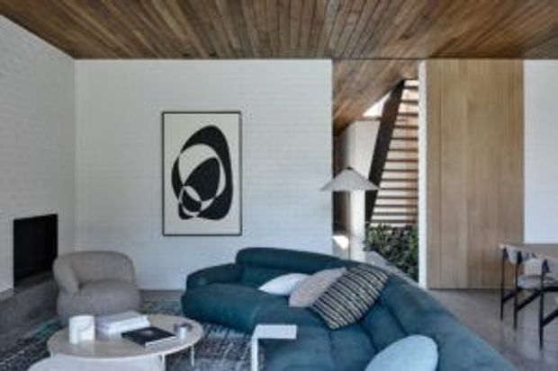 Contemporary living room with a large curved blue sectional sofa, sculptural lounge chair, round coffee table, and abstract monochrome artwork. The room features white walls, a warm timber-lined ceiling, extensive timber wall panelling, and a partially visible open stair with horizontal timber treads or slats.