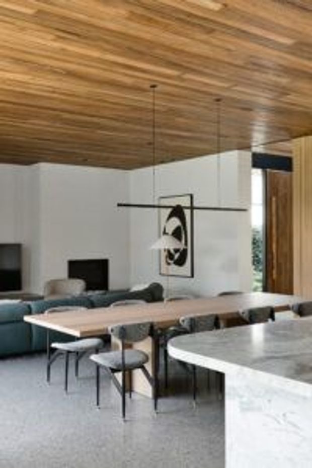 Open-plan contemporary living and dining area with a warm timber-lined ceiling, white walls, a long light-wood dining table, dark upholstered dining chairs, a teal sofa, and a curved pale stone kitchen island in the foreground. A minimalist linear pendant light hangs over the dining table, with timber-framed glazing or doors at the rear.