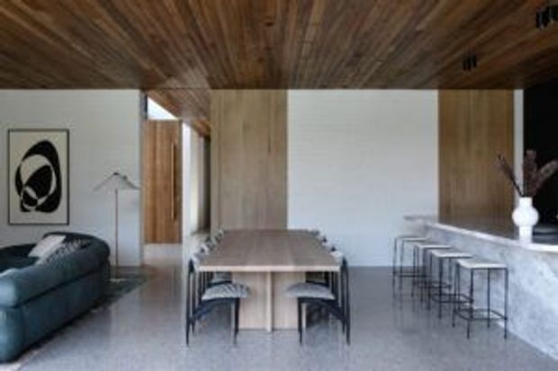 Open-plan contemporary dining and living area with a long light-wood dining table, upholstered dining chairs, a dark curved sofa, and a stone-topped island or counter with bar stools. The room features extensive warm timber ceiling and wall panelling, white walls, terrazzo-like flooring, and minimalist artwork and styling.