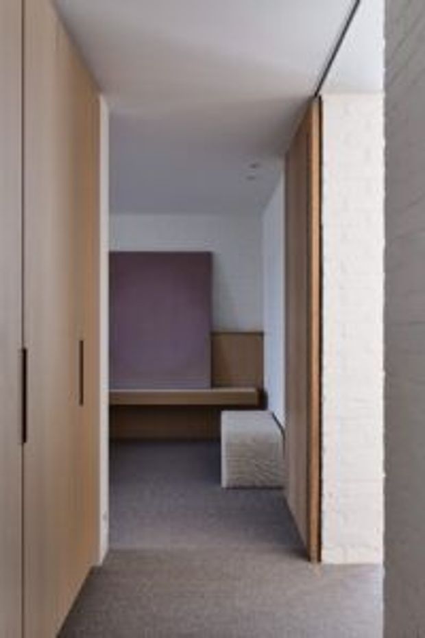 A narrow, minimalist interior passage framed by tall light timber cabinetry and door panels, leading into a compact bedroom or study nook. The space has pale walls and ceiling, grey carpet, integrated timber joinery, a built-in desk or bench, and a muted mauve upholstered or painted feature panel at the far end. A small light upholstered cube sits beneath the joinery.