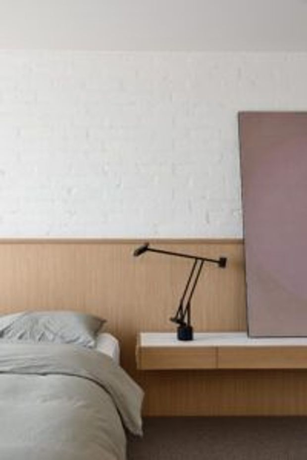 Close-up of a contemporary bedroom featuring a partially visible bed with light grey bedding, a timber wall panel and integrated floating bedside shelf, a slim black adjustable wall-mounted reading lamp, and a large muted mauve artwork or panel. The palette is soft and neutral with warm timber detailing and dark carpet.