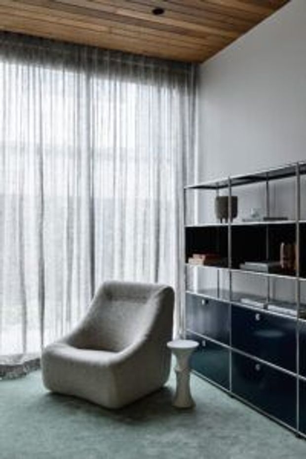 A refined modern living room corner featuring a sculptural upholstered lounge chair and small side table beside a dark modular shelving and storage unit. Full-height sheer curtains diffuse daylight across the room, while a warm timber slatted ceiling contrasts with the pale walls and muted grey carpet.