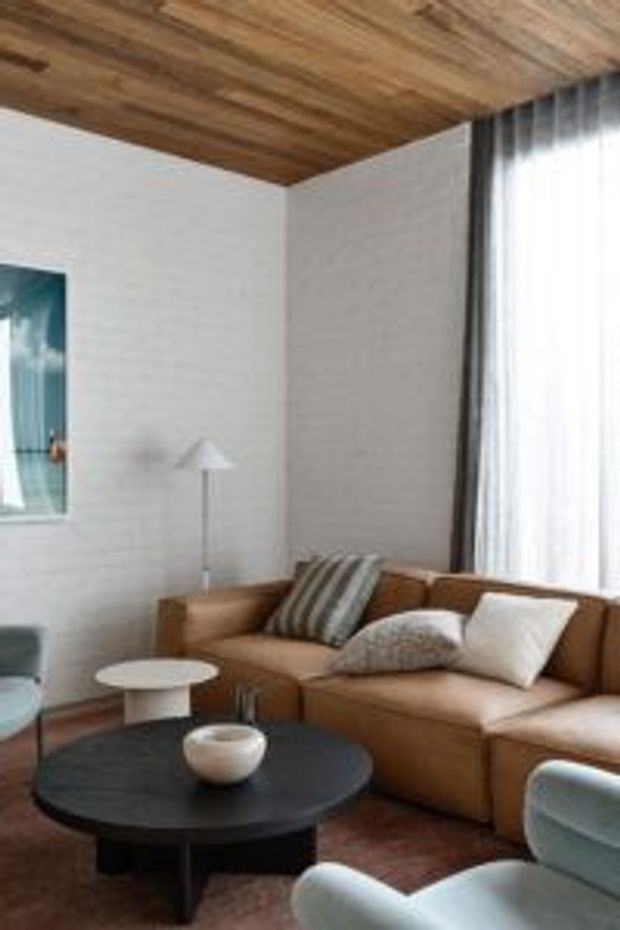 A warm, refined living room with a large tan sectional sofa, sculptural coffee tables, upholstered lounge chairs, sheer floor-to-ceiling curtains, white textured walls, and a striking timber-lined ceiling. The space combines natural materials with muted contemporary furnishings and soft daylight.