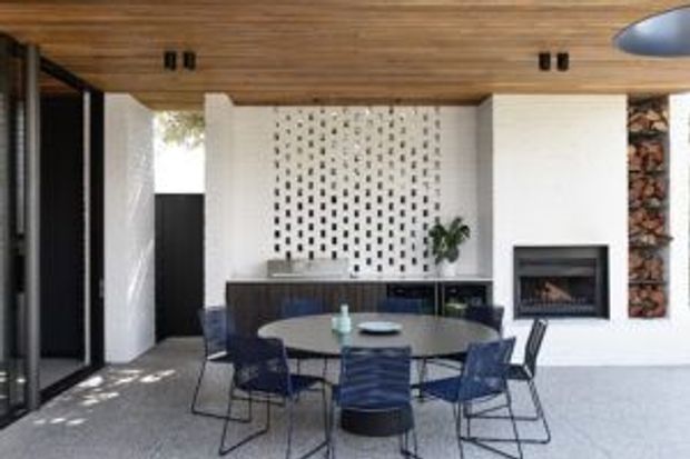 Contemporary open-plan dining area with a round dark dining table and blue upholstered chairs, positioned beside a compact kitchen with dark cabinetry. A white feature wall incorporates a patterned breeze-block screen, fireplace, and timber-lined ceiling. Full-height black-framed glazing opens to the exterior, creating a bright indoor-outdoor connection.