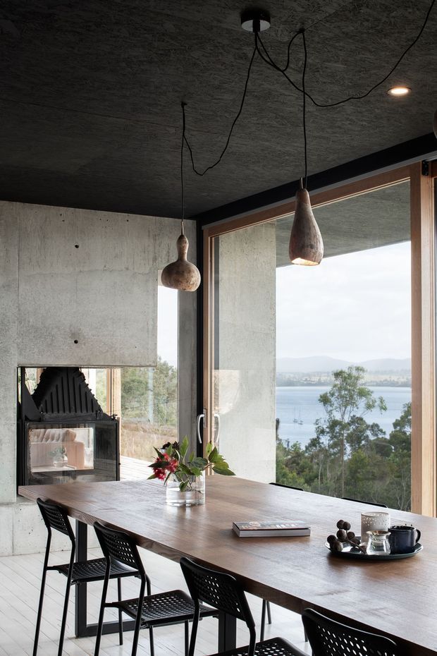 A moody contemporary dining area with a long dark timber dining table and black woven chairs, positioned beside expansive timber-framed glazing overlooking a body of water and distant hills. The room features raw concrete walls and ceiling, a black-framed fireplace, and sculptural pendant lights with exposed cords. A floral arrangement, books, and tableware provide restrained decorative accents.