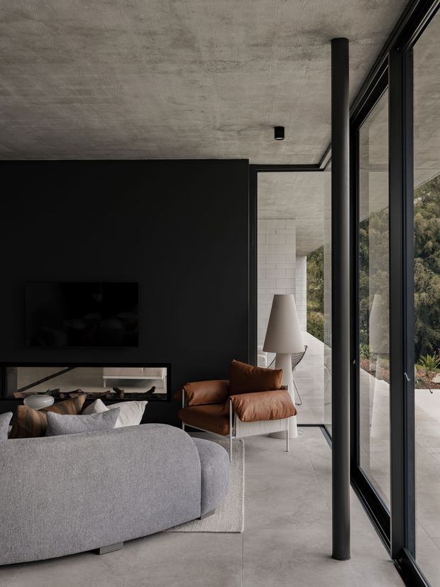 A dark, refined contemporary living room viewed from beside full-height black-framed glazing. The room features a charcoal feature wall with a wall-mounted television and low horizontal fireplace or display opening, a large curved light-grey sofa, a brown leather lounge chair, a sculptural floor lamp, and pale stone or concrete-look flooring. The exposed textured concrete ceiling, black cylindrical column, and extensive glazing create a minimalist indoor-outdoor connection, with landscaped surroundings visible beyond the windows.