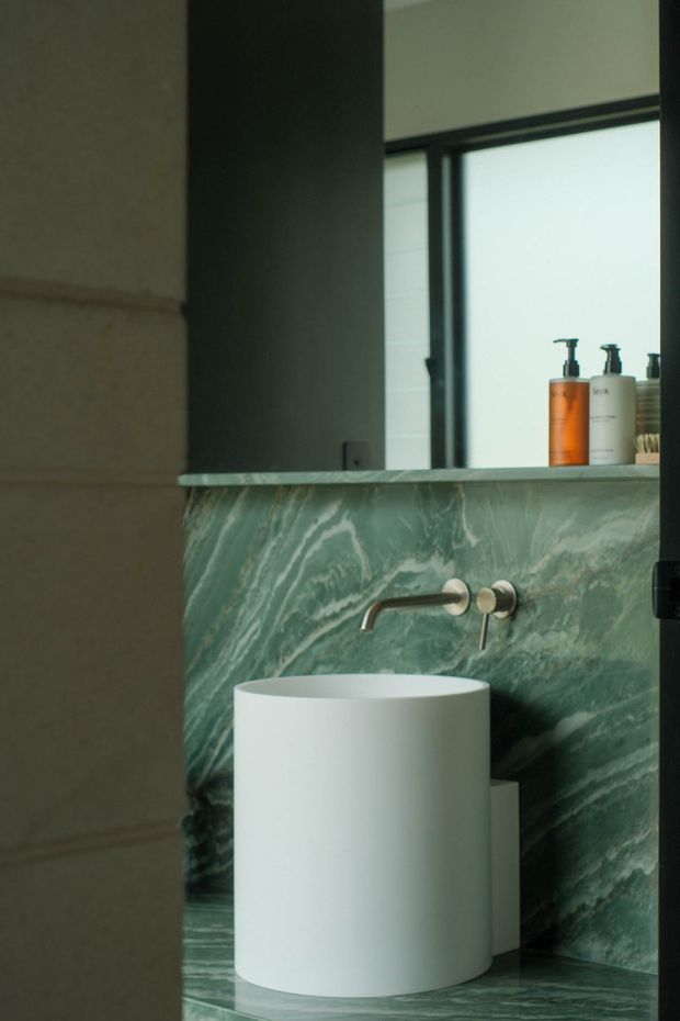 Close-up view of a refined bathroom vanity featuring a cylindrical white vessel basin, wall-mounted brushed metal tapware, and dramatic green veined stone cladding across the vanity wall and benchtop. A large mirror, dark-framed window, and toiletries are visible above the basin. The space has a moody, sophisticated contemporary finish.