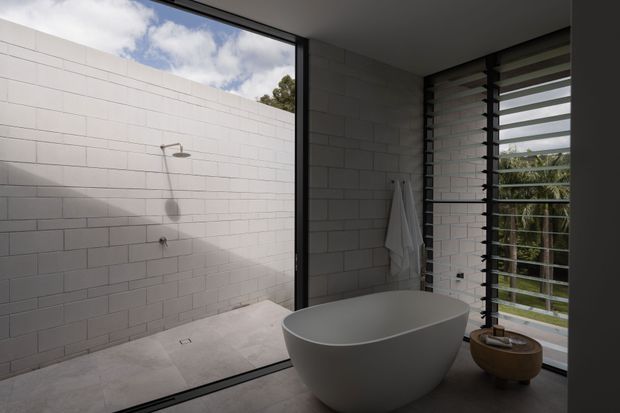Contemporary bathroom with a freestanding white oval bathtub beside full-height louvred glazing. The open walk-in shower is enclosed by pale rectangular masonry-style wall tiles and matching floor tiles, with dark window framing and views to greenery outside. The room has a calm, light-filled, minimalist character.
