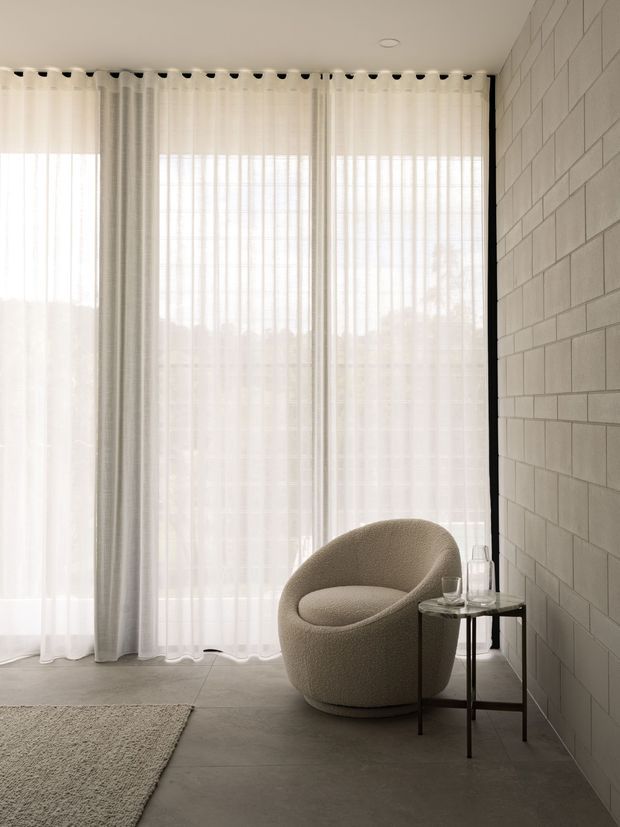A calm, minimalist interior corner with a curved upholstered lounge chair beside a small dark side table. Full-height sheer curtains diffuse daylight across a tiled or concrete-look floor, while a pale exposed-block feature wall adds texture. A neutral rug is partially visible in the foreground.