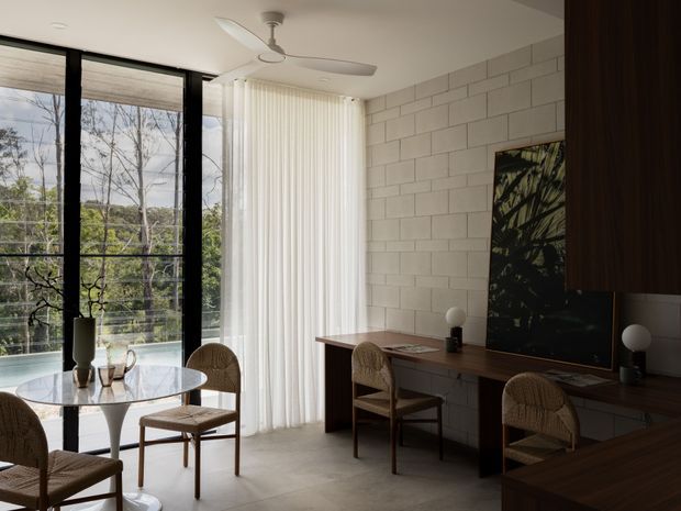A refined contemporary dining and work area with a small round glass dining table and woven cane chairs beside full-height black-framed glazing. Sheer vertical curtains soften the daylight and leafy outdoor views. A long dark timber console or desk runs along a white painted masonry feature wall, with sculptural table lamps and artwork. The space has polished concrete flooring, a ceiling fan, and a warm, restrained palette.