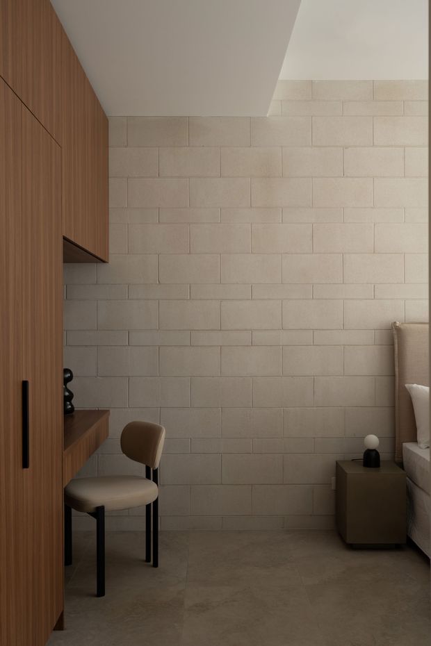 A warm, minimalist bedroom corner with a compact built-in timber desk and cabinetry along the left wall, a softly upholstered chair, a stone-block feature wall, and a partially visible bed with a dark bedside table on the right. The restrained palette and concealed joinery create a calm, refined atmosphere.
