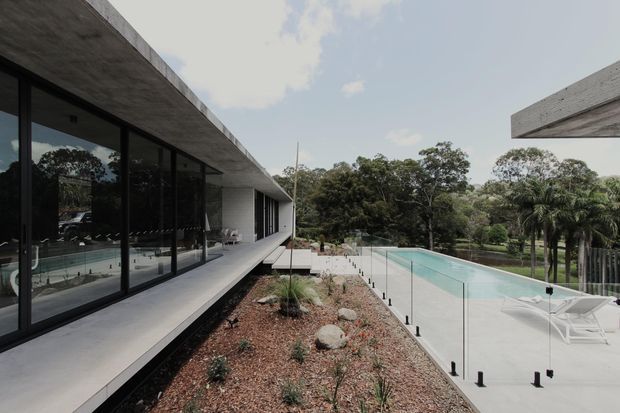 Contemporary low-profile residence with broad cantilevered concrete roof planes, dark floor-to-ceiling glazing, and a long elevated terrace overlooking a minimalist swimming pool. The landscaped garden uses gravel, rocks, and sparse planting, while glass pool fencing and pale concrete create a clean modern outdoor setting.