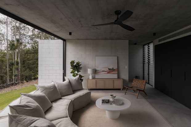 A refined contemporary living room with a large curved sectional sofa, round coffee table, timber sideboard, lounge chair, and abstract artwork. The room features exposed raw concrete ceilings, white painted brick walls, extensive floor-to-ceiling glazing, dark built-in cabinetry, and a ceiling fan. It opens directly to a landscaped garden and wooded setting.