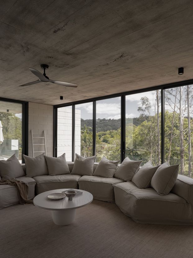 A calm, contemporary living room with a large low-profile sectional sofa arranged around a round coffee table. Floor-to-ceiling black-framed glazing wraps the room, overlooking a wooded landscape. The interior features a raw-looking concrete ceiling, pale neutral upholstery and flooring, a minimalist ceiling fan, and a white block wall with a ladder detail.