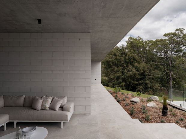 A minimalist covered outdoor lounge beside a white masonry wall, with a low upholstered sofa and round coffee table on a pale concrete floor. A dramatic cantilevered concrete roof creates a sheltered terrace overlooking landscaped bushland and a partially visible pool area. The composition is quiet and highly restrained, with strong geometric lines and integrated indoor-outdoor connection.