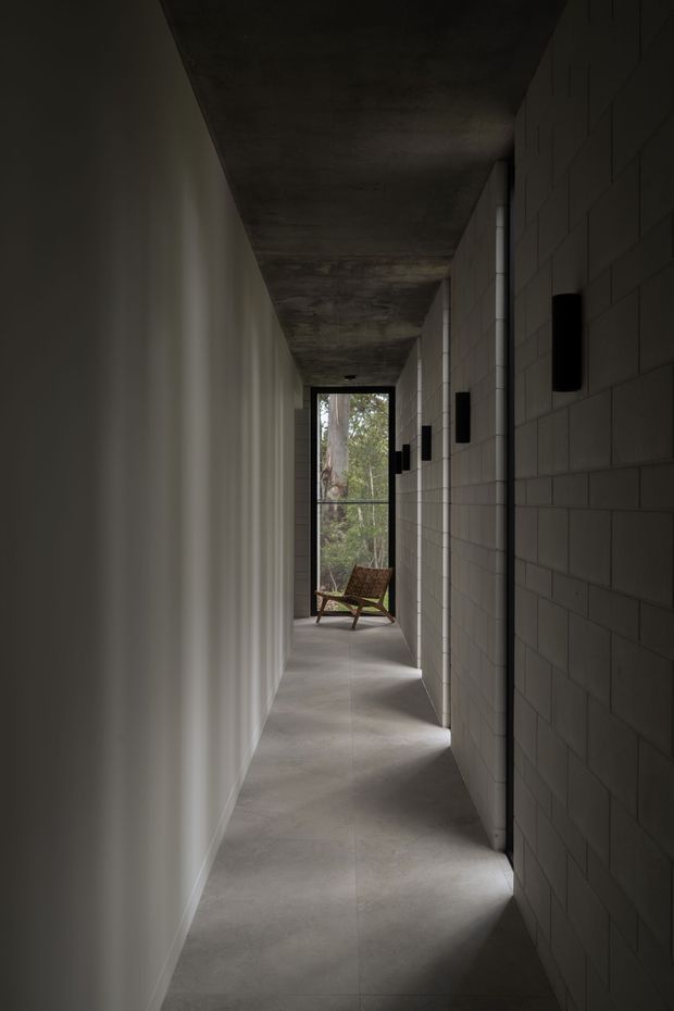A long, narrow architectural corridor with a raw concrete ceiling and polished grey concrete floor. One side has smooth pale walls with deep vertical recesses, while the other is formed from exposed grey concrete blockwork with black wall-mounted light fixtures. Natural light enters through a full-height glazed opening at the far end, where a wooden lounge chair is positioned, creating a quiet, restrained atmosphere.