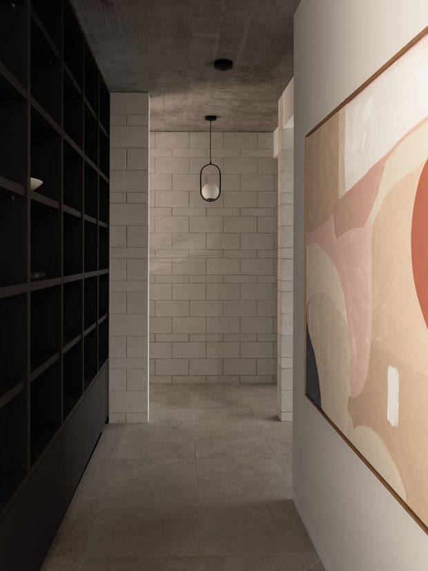 A narrow, dimly lit interior passage with a raw industrial character. The space has a polished concrete ceiling and large-format concrete-look flooring, a full-height black built-in shelving unit along the left wall, and a white-painted wall on the right displaying a large abstract artwork in a timber frame. At the end, a recessed alcove is lined with light grey rectangular masonry or tile blocks and illuminated by a simple suspended pendant light.