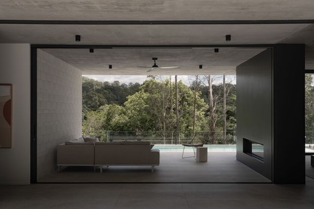 A dramatic contemporary living room with a low concrete ceiling, dark-framed full-height sliding glazing, pale blockwork walls, and a minimalist fireplace wall. The room opens directly onto a terrace and pool overlooking dense bushland, creating a strong indoor–outdoor connection. A neutral sofa, small table, lounge chair, and ceiling fan furnish the space, while recessed spotlights and dark framing add contrast.