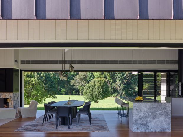 Open-plan contemporary living and dining area with a large retractable glazed opening to a landscaped garden. The room features timber flooring, a dark dining table with upholstered chairs, a patterned area rug, a pale stone kitchen island, dark cabinetry, extensive black-framed glazing, and a striking vertically ribbed ceiling or soffit detail. Pendant lighting hangs over the dining table.