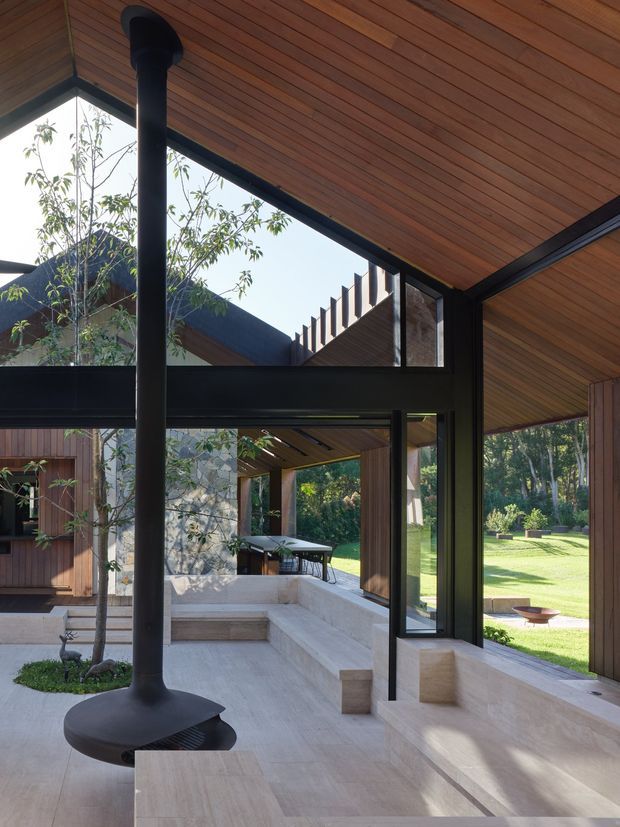 A light-filled contemporary living area with expansive black-framed glazing opening onto a landscaped garden courtyard. The space features a dramatic freestanding black suspended fireplace, pale stone or concrete floors and built-in seating, warm timber-lined ceilings and walls, and strong indoor-outdoor connections. A partial view of an outdoor dining area and adjacent timber-clad structure is visible beyond the glazing.