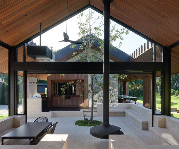 A contemporary open-plan living and dining pavilion with a dramatic pitched timber-lined ceiling, black-framed floor-to-ceiling glazing, and a central suspended fireplace flue with a circular hearth. The space opens onto a landscaped courtyard and lawn, with built-in pale stone seating, an outdoor kitchen or bar area, and a timber-clad structure visible beyond. The interior has strong indoor-outdoor connectivity and abundant natural light.