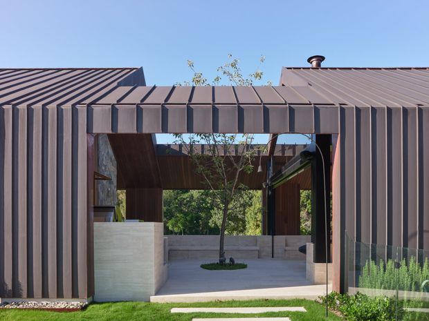 Contemporary residential exterior featuring a sculptural metal-clad gateway or covered passage between low building wings. The dark standing-seam and vertically ribbed metal cladding forms a strong geometric frame, opening onto a landscaped internal courtyard with a young tree, pale concrete walls, paving, and timber-lined soffits. Clear blue sky and clipped lawn surround the structure.