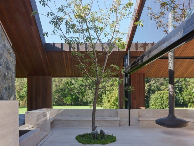 Open-air covered architectural courtyard or living space with warm timber-lined soffits, exposed structural beams, stone-clad walls, pale stone seating and paving, and a mature tree planted through an opening in the roof. Large glazed openings connect the space to a lush garden, while a sculptural dark pendant light hangs beneath the canopy. The design has a strong indoor-outdoor connection and a calm, refined natural character.