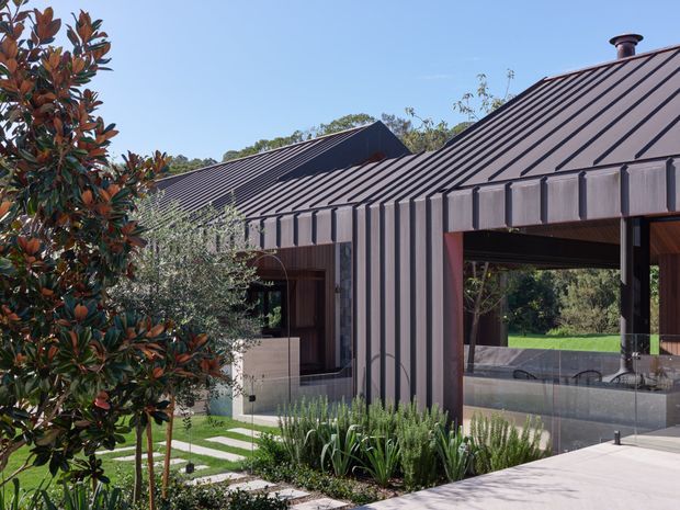 A contemporary single-storey residential building set within landscaped grounds. The structure features dark standing-seam metal cladding and pitched roofs, large glazed openings, timber-framed outdoor living areas, and a pale concrete terrace with planted garden beds. A clear, sunny sky and mature planting surround the modern low-profile form.