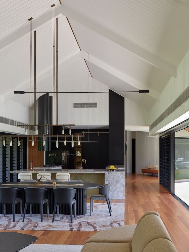 A sophisticated open-plan kitchen, dining and living space beneath a dramatic white pitched, timber-lined ceiling. The kitchen features a large stone island with brass tapware, dark built-in cabinetry, a long dining table with black upholstered chairs, and sculptural linear pendant lighting. Warm timber flooring, a patterned area rug, black-framed glazing and a neutral sofa create a refined contemporary interior with strong contrast between light architectural surfaces and dark joinery.
