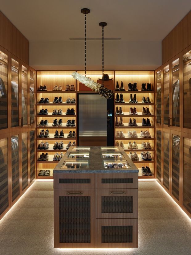 A compact, highly organized walk-in shoe wardrobe or dressing retail-style display room. Warm timber wall-to-wall cabinetry with illuminated open shelves and glass-fronted side cupboards displays numerous pairs of shoes. A central timber island with glass display panels and integrated ventilation grilles occupies the room, while concealed LED strip lighting outlines the shelving, cabinetry bases, and island. Two dark chain-hung pendant lights and a decorative suspended branch-like feature hang from the plain ceiling. The space has a luxurious, warm, bespoke joinery aesthetic.