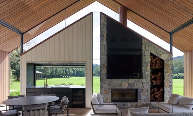 A bright, open-plan living and dining space beneath a dramatically pitched timber-lined ceiling. The room features a central stone-clad fireplace and television wall with integrated firewood storage, a contemporary kitchen visible through a large opening, and full-height glazing overlooking expansive green grounds. Dark-framed structural glazing and restrained modern furniture create a refined indoor-outdoor connection.