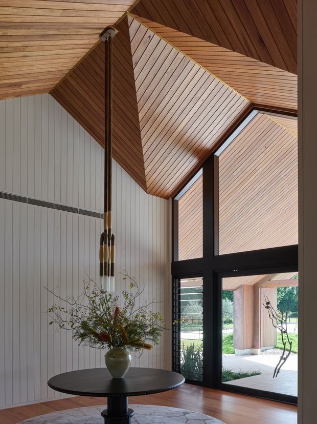 Interior view of a refined contemporary entry or living space with a dramatic angular timber-lined vaulted ceiling, white vertical wall panelling, dark-framed floor-to-ceiling glazing, and a round dark timber table with a sculptural floral arrangement. The glazing looks onto a landscaped outdoor area and a covered timber-clad structure. The space has strong geometric ceiling planes, warm natural timber, and abundant daylight.