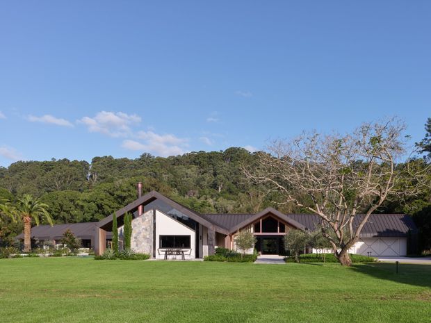 Single-storey contemporary residence set in a broad manicured lawn, with multiple steeply pitched gabled roof forms, dark metal roofing, stone-clad feature walls, timber-toned framing and extensive glazing. The home is surrounded by mature trees and landscaped planting under a clear blue sky.