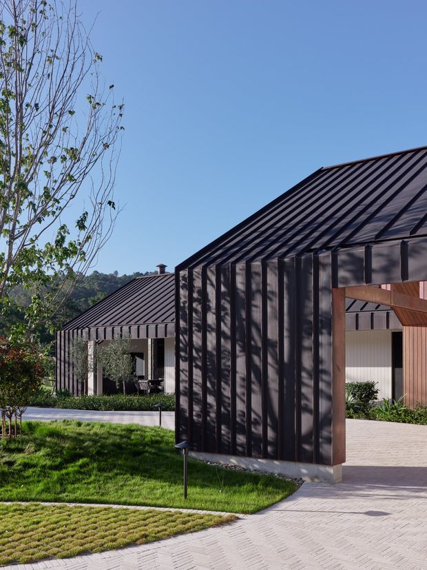 Exterior architectural photograph of a contemporary property composed of dark ribbed metal-clad pavilion-like structures with steep pitched roofs. A large foreground volume frames a covered entry or passage with warm timber detailing, while a matching smaller building sits beyond landscaped lawns and paving. Clear blue sky, mature planting, and strong tree shadows create a crisp, modern resort-like setting.