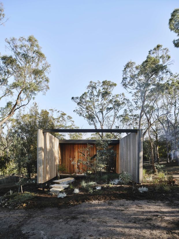 A small contemporary pavilion or cabin set within a native eucalyptus woodland. The open-fronted structure has tall vertical timber-clad side walls, a dark flat roof frame, and a warm timber rear wall with glazed or recessed openings. Stone stepping slabs lead through a landscaped garden of low native planting, rocks, and mulch.